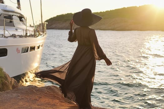 woman in long, flowing dark dress and a wide-brimmed hat standing on a rocky shore next to a white yacht during sunset.