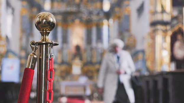 Golden barrier pole with red rope and chain limiting access inside a church with blurred tourist walking