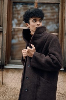 Fashion photo of a young man in a brown coat standing in front of a window, with hands in pockets and head tilted to the side.