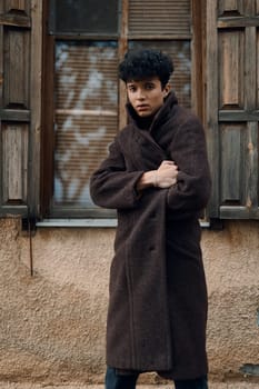 Fashionable young man in a brown coat posing in front of an old building, with hands on hips and looking at the camera.