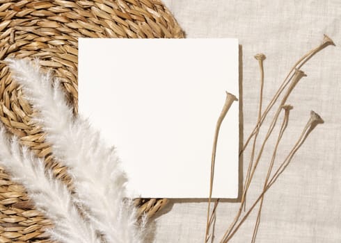 Blank card near dryed grass on beige vowen placemat and linen fabric top view with hard light, copy space. Boho flat lay with square card Bohemian or close to nature wedding stationery