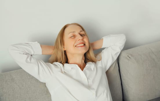 Happy modern relaxed young woman enjoying at home, satisfied girl resting with closed eyes and folded hands behind the head sitting on the couch in living room