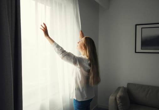 Woman stands by the window, opening curtains at home in morning, household chores, routine or interior decoration after renovation, side view