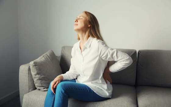 Woman suffering from back pain, touches her lower back while sitting on couch in living room at home