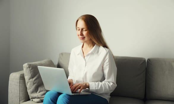 Happy modern woman working with laptop, freelancer girl looking at computer sitting on a couch in the living room at home