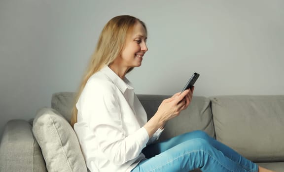 Happy smiling mature woman looking at phone, girl with smartphone sitting on the couch in living room at home