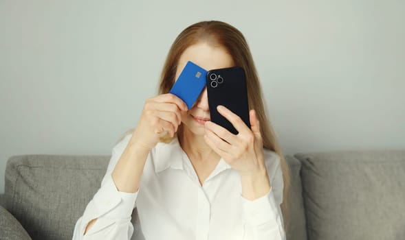 Upset modern young woman holding smartphone and credit card sitting on the couch at home, internet fraud, problems with online payments or blocked bank card