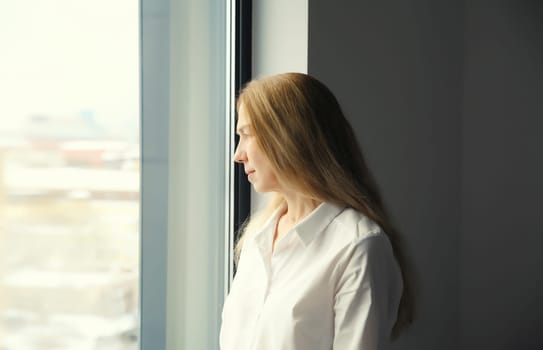 Pensive young woman thinks looks out the window, thoughtful girl suffering from depression standing in living room at home
