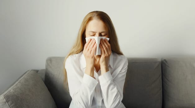 Sick exhausted young woman sneezing blow nose using tissue, unhealthy suffering girl feels unwell sitting on the couch at home