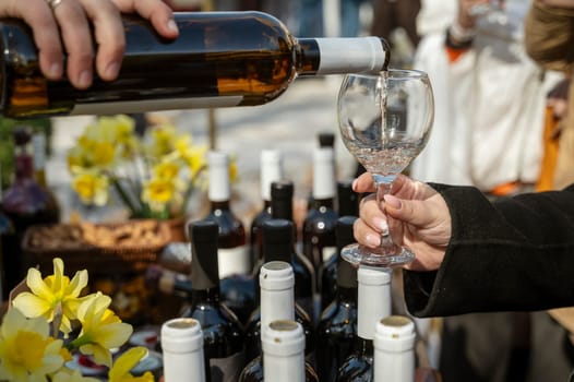 Close-up of a person's hand pouring white wine into a glass, many bottles of wine nearby. Yellow flowers in the background, shallow depth of field. The image conveys the elegance of a fine dining experience.