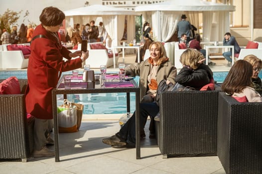 Odessa, Ukraine - April 05, 2025: City food festival, group of middle aged women sitting at table and taking photos on phoneand drinking wine. table near swimming pool
