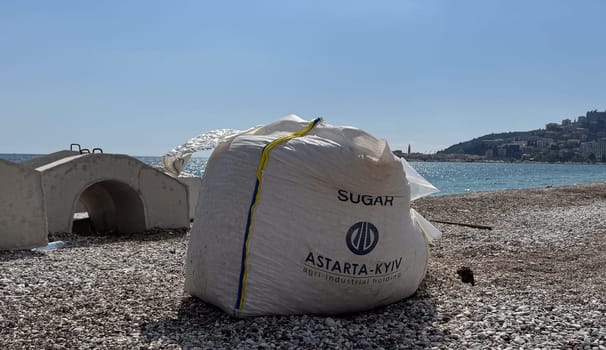 Budva, Montenegro - April 17, 2025: A bag with the inscription SUGAR ASTARTA KYIV, with pebbles and stones inside, stands next to the reinforcing structure of the embankment