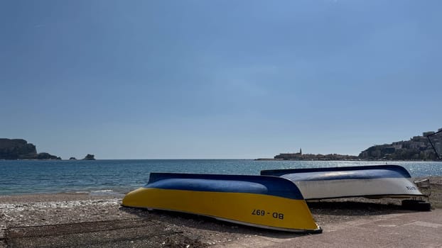 Budva, Montenegro - April 17, 2025: Colorful boats stand on a sandy beach with a serene sea and the Old Town in the distance