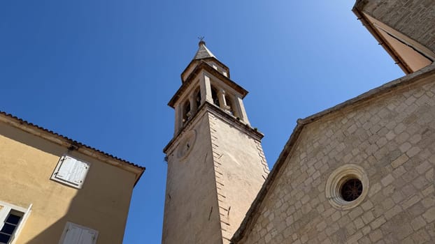 Budva, Montenegro - April 20, 2025: The historic Church of St. John the Baptist with a church tower rises against a clear blue sky