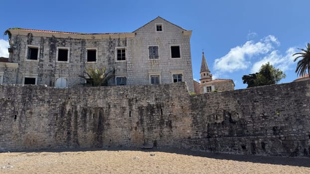 Budva, Montenegro - April 18, 2025: An abandoned historic building located on the seafront, with palm trees and a church tower, embodies history and the passage of time.