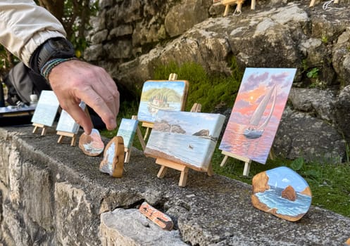 Budva, Montenegro - April 18, 2025: Artist's hand arranges painted stones and paintings displayed on the shore, a concept of artistic expression