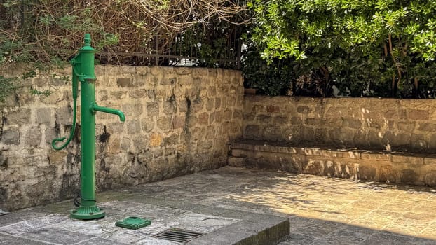 Budva, Montenegro - April 18, 2025: An old green water pump stands in the old town next to the La Mia Pasta restaurant, surrounded by lush greenery, stone walls and benches.