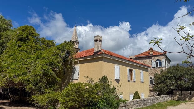 Budva, Montenegro - April 18, 2025: Budva, Montenegro - April 18, 2025: Historic Mediterranean villa in the old town, surrounded by vibrant greenery against a clear blue sky