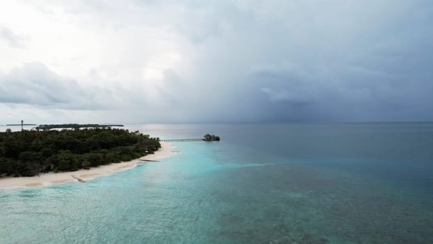 Breathtaking aerial view of a tropical island in the maldives, showcasing crystal-clear turquoise waters, lush greenery, and an overwater bungalow under a cloudy sky