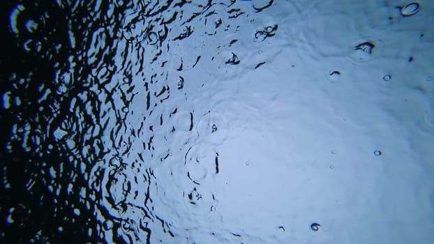 Rain falling on a blue water surface creates concentric circles, distorting the reflection of a dark object and forming an abstract, textured effect in nature