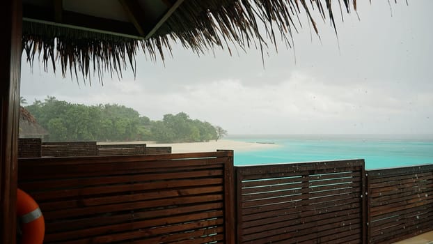 Relaxing in a tropical beach hut with a thatched roof, enjoying the stunning view of a turquoise ocean under a refreshing rain shower, surrounded by lush greenery and tranquil skies