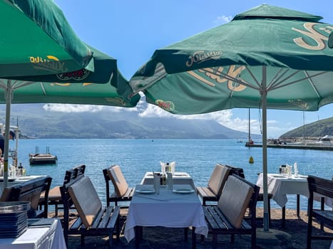 Budva, Montenegro - April 18, 2025: A picturesque waterfront restaurant with green umbrellas and breathtaking mountain views creates a serene atmosphere in which guests enjoy their meals