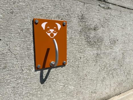 Metal hook for leashing dogs on textured concrete wall, part of urban infrastructure supporting pet friendly environments in public spaces. Bright plate with dog symbol offers clear purpose
