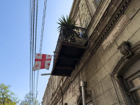 Old balcony with iron railing and Georgian flag waving against blue sky. National identity symbol on weathered building facade in Tbilisi historical district, reflecting cultural heritage