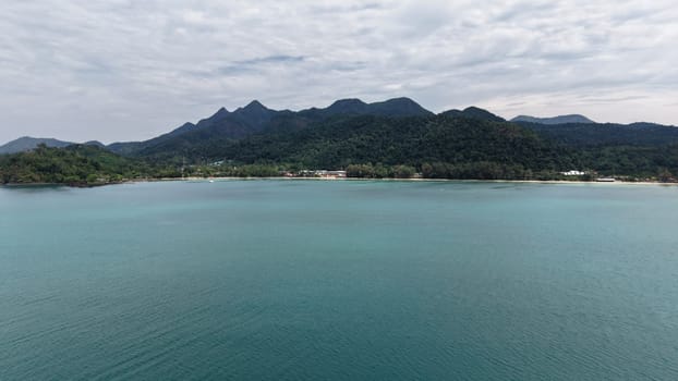 Turquoise water gently laps against the sandy shore of a tropical island, with lush green mountains rising majestically in the background under a cloudy sky, creating a serene and picturesque scene
