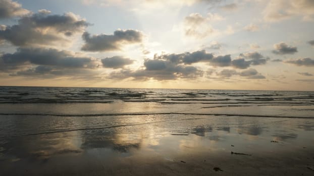 Golden sunset reflecting on wet beach sand with waves gently crashing on the shore and a cloudy sky creating a serene and peaceful atmosphere