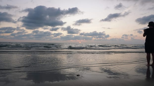 Photographer standing on a wet sandy beach at sunset, capturing vibrant sky colors reflected in gentle waves as the sun sets below the horizon