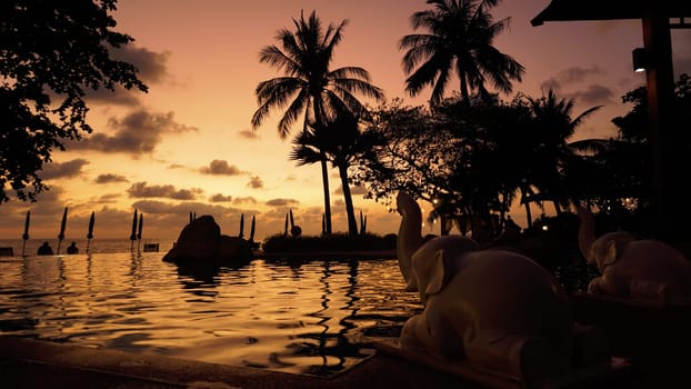Two elephant statues are placed at the edge of an infinity pool during a golden sunset in a tropical resort, with palm trees and beach umbrellas silhouetted against the colorful sky