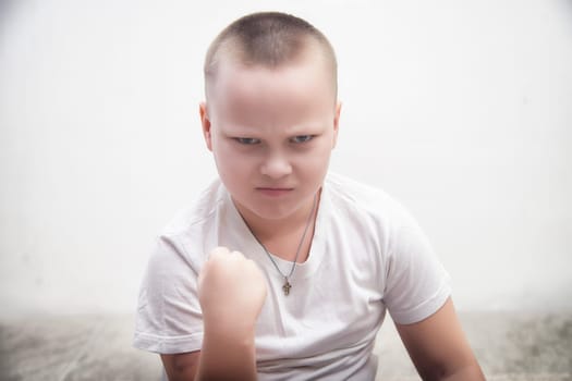 A boy is expressing strong emotions with a clenched fist, wearing a plain white shirt, in a simple room with white walls.