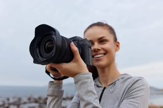A smiling young woman in a gray athletic jacket holds a professional camera at the beach Her joyful expression captures the essence of creativity and outdoor photography