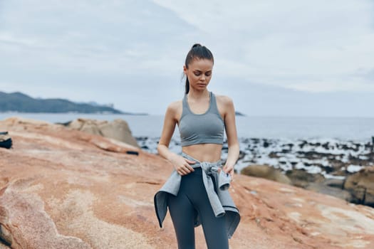 Fit female model in gray sports clothing poses on rocky shoreline, embodying healthy lifestyle and fitness with a calm ocean backdrop, inspiring a sense of well being