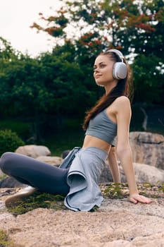 Relaxed young woman in athletic wear with headphones, sitting on a rock in a green outdoor setting, promoting a healthy lifestyle and enjoyment of music