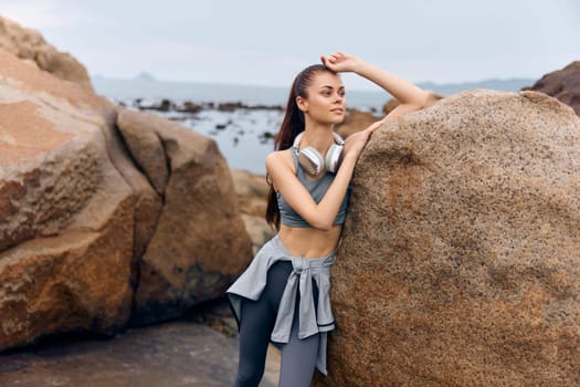 Confident young woman in athleisure wear standing by a rocky beach, promoting a healthy lifestyle and fitness, with headphones around her neck, enjoying her workout