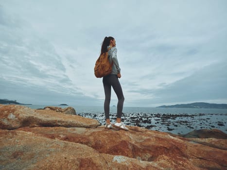 A young woman with a backpack standing on a rocky shore, dressed in athletic wear, embodying exploration and freedom by the ocean, perfect for travel and adventure themes