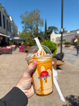 Tbilisi, Georgia, April 11, 2025: Iced coffee with ice cream in branded Tolia cup held outdoors on sunny day, takeaway drink with caramel topping on lively pedestrian street with cafes and benches