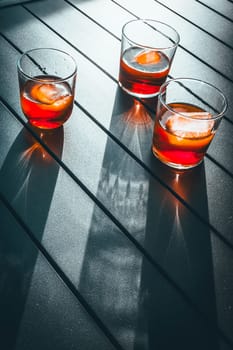 Artistic image of three glasses of red vermouth with ice casting long shadows on a metallic terrace table in bright sunlight. The image captures a Mediterranean lifestyle moment of enjoying a sunny aperitif