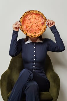 Woman sitting on a green chair and holding a pizza in front of her face, hiding her identity with a playful gesture