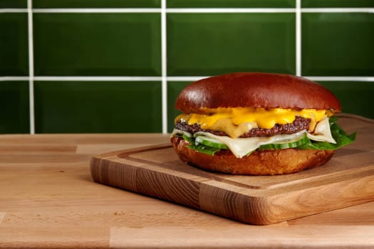 Tasty cheeseburger with melted cheese, pickles, and lettuce served on a wooden cutting board in a contemporary kitchen with green tiles
