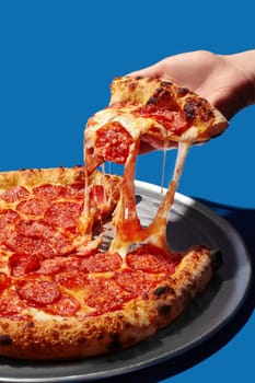 Hand lifting a slice of hot pepperoni pizza, with stringy melted cheese against a vibrant blue background