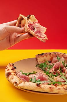 Hand holds a slice of delicious pizza with prosciutto and arugula over a whole pizza on a plate with a vibrant, colorful background