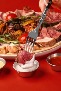 Woman using a fork dipping grilled beef into white sauce, enjoying a variety of grilled meat and vegetables