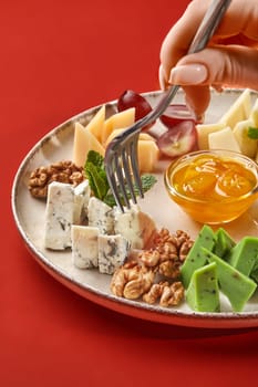 Close-up of a woman using a fork taking a piece of gorgonzola cheese from a plate with assorted cheeses, walnuts, grapes, and honey