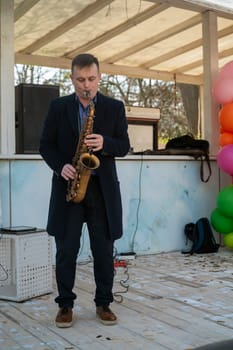 Odessa, Ukraine - April 05, 2025: City food fair. A man in a coat plays the saxophone, on a makeshift stage made of white wood and with colorful balloons.