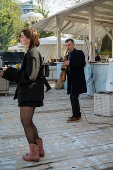 Odessa, Ukraine - April 05, 2025: City food fair. Man in coat plays saxophone, young stylish girl in dark jacket and boots dances nearby.