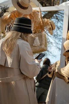 Portrait of a young girl in a beige trench coat and a black hat, seen from the back. She is standing in front of a mirror in front of a street stall choosing a hat and talking on the phone.