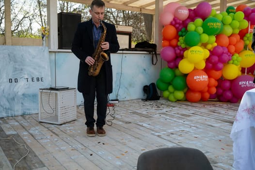 Odessa, Ukraine - April 05, 2025: City food fair. A man in a coat plays the saxophone, on a makeshift stage made of white wood and with colorful balloons